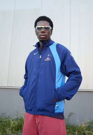 Navy and light blue windbreaker jacket with logo and zip pockets. Features elastic cuffs and a stand-up collar. Worn over red shorts.