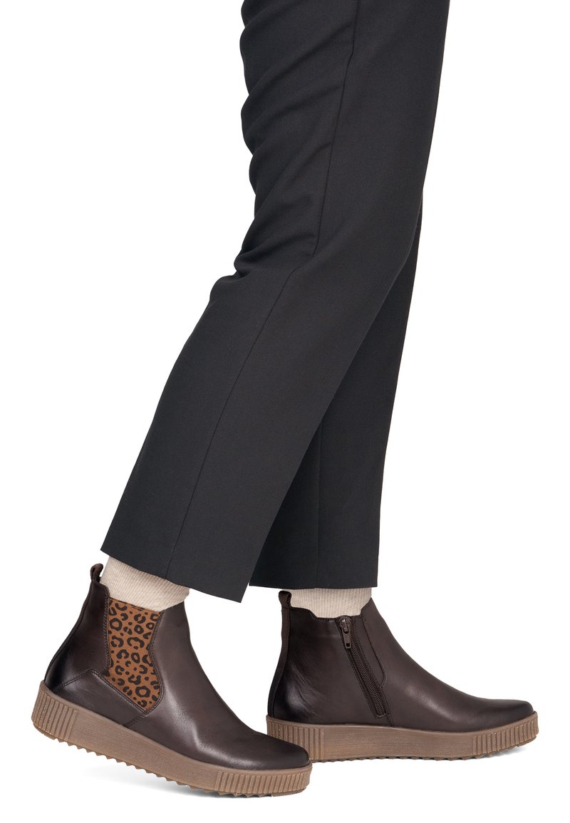 Personne portant un pantalon noir, des chaussettes beiges et des bottines marron foncé avec un panneau élastique imprimé léopard et une fermeture éclair latérale, debout les pieds croisés.