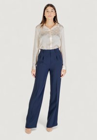 Blouse en tissu crémeux transparent avec un motif subtil et des boutons, associée à un pantalon taille haute bleu marine avec des plis et une coupe ajustée.