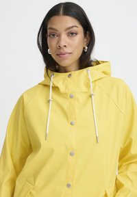 Veste imperméable jaune avec capuche, ornée de cordons blancs et de boutons-pression argentés. Texture lisse et coupe décontractée.