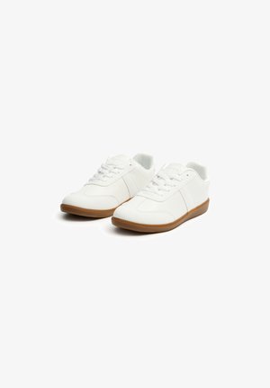 Zapatillas blancas hechas de material sintético con suela de goma texturizada. Incluyen características como puntera redonda, cordones y dos franjas laterales.