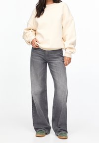 Przesadny kremowy sweatshirt, szare dżinsy o szerokich nogawkach, zielone sneakersy z jasnobrązowymi podeszwami. Miękka tkanina, luźny krój, ściągacze u rękawów i na dekolcie.