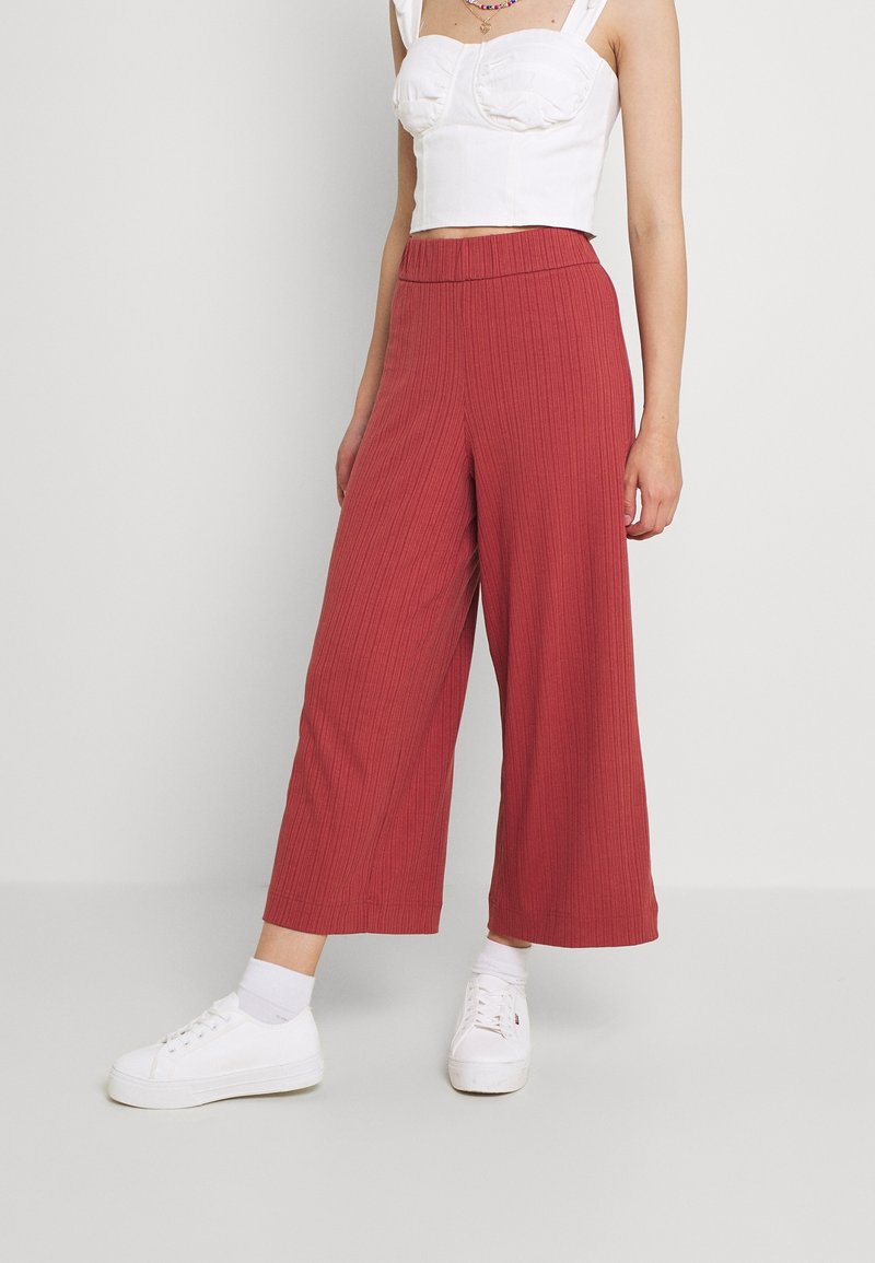 Femme portant un pantalon taille haute, large, de couleur rouille avec un crop top blanc, des chaussettes blanches et des baskets compensées blanches sur un fond uni.