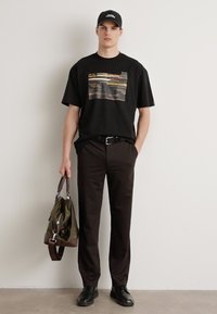 Svart t-skjorte med flerfarget grafisk design, svarte bukser, svarte sko og en olivengrønn bag med brune detaljer.