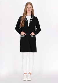 Schwarze Strickjacke mit weißen Streifenakzenten, langen Ärmeln, Knopfverschluss und Fronttaschen. Darüber ein weißes Oberteil getragen und kombiniert mit weißen Hosen.