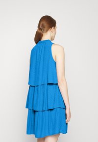 Vestido azul em camadas com gola alta, design sem mangas, tecido texturizado e detalhes franzidos, apresentando um padrão de bolinhas.