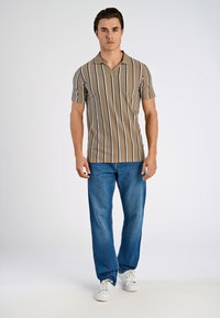 Beige kortärmad polo skjorta med vertikala svarta och vita ränder, i kombination med blå denim jeans och vita sneakers, stående framför en enkel bakgrund.