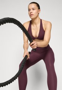 Athletic outfit in deep burgundy, featuring a halter-style top and fitted leggings. Black battle rope being held tightly, textured grip visible.