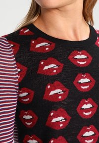 Strickpullover mit auffälligem Muster roter Lippen auf schwarzem Hintergrund, mit gestreiften Ärmeln in Rot und Weiß. Rundhalsausschnitt.