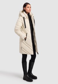 Parka in crema con cuciture orizzontali, cappuccio removibile e dettagli in metallo neri. Il modello indossa leggings neri e stivali al ginocchio.