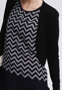 Cardigan nero sopra un vestito blu navy con motivo a zigzag bianco. Il vestito presenta uno scollo rotondo e una vestibilità ampia, con una texture morbida.