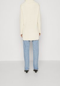 Eine stehende Person trägt einen langen cremefarbenen Strickpullover, helle blaue gerade Jeans und schwarze High Heels vor einem schlichten weißen Hintergrund.