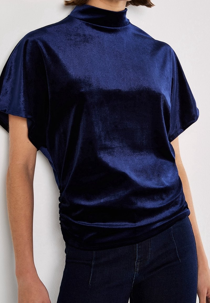 Femme portant un top en velours bleu foncé à manches courtes et col montant, ainsi qu’un pantalon foncé, sur un fond uni.