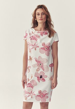 Frau mit hellbraunem Haar trägt ein weißes knielanges Kleid mit großem rosa Blumenmuster und steht vor hellem, einfarbigem Hintergrund.