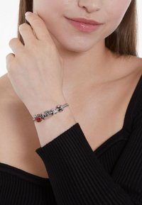 Zilveren armband met kleurrijke bedels, waaronder een rode kraal en metalen accenten. Draagt om een pols, gecombineerd met een zwart geribd topje.