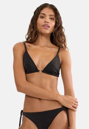 Frau trägt schwarzen glitzernden Bikini mit Schnürung und Goldringdetail, posiert mit einem Arm über der Taille auf weißem Hintergrund.