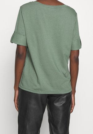Persoon draagt een losse groene korte mouwen shirt met opgerolde manchetten en zwarte leren broek, van achteren gezien tegen een witte achtergrond.