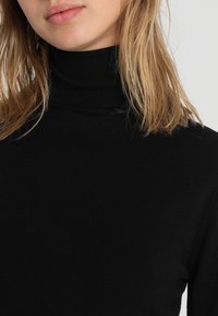 Blond person som bär en svart ribbad polotröja, där endast läppar, haka, hals och överkropp syns mot en enfärgad bakgrund.