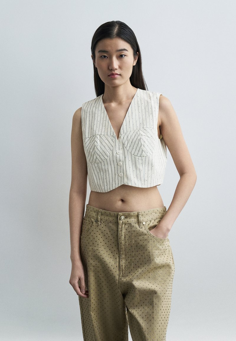 Νεαρή γυναίκα με μακριά σκούρα μαλλιά φοράει ένα αμάνικο ριγέ crop top και μπεζ παντελόνι με ψηλή μέση και μικρά σχέδια σε μορφή κουκίδων, με το χέρι στην τσέπη.