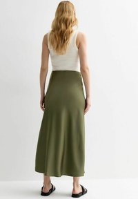 Falda larga de satén color verde oliva con una textura suave, que presenta una cintura ajustada y un diseño fluido. Combinada con una camiseta sin mangas blanca y sandalias negras.