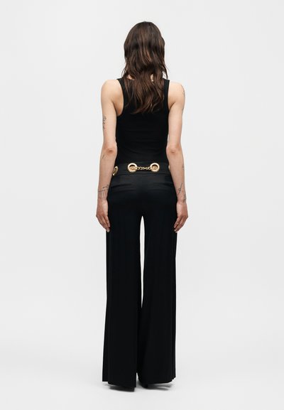 Γυναίκα με μακριά καστανά μαλλιά που κοιτάζει από την πλάτη, φοράει μαύρο αμάνικο τοπ, φαρδιά μαύρα παντελόνια και μια χρυσή αλυσίδα ζώνης.