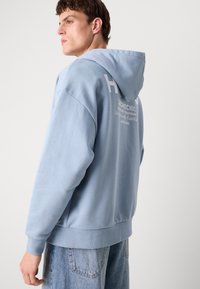 Lichtblauwe hoodie met een textuurafwerking, uitgerust met een grote capuchon, geribbelde manchetten en bedrukte logodetails op de achterkant. Gecombineerd met lichte denim.