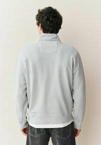 Lysegrå sweatshirt med høj krave og ribbede manchetter, med en rektangulær lap på ryggen og blød tekstur. Båret over lagdelte skjorter.