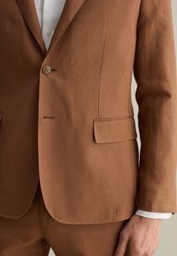 Blazer in lino marrone con chiusura a un bottone, una tasca sul petto e due tasche laterali. Tessuto liscio con una vestibilità sartoriale.