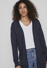 Dunkelblaue, offene Strickjacke mit langen Ärmeln, aus einem weichen Material gefertigt. Verfügt über zwei Fronttaschen und eine lockere Passform. Wird über einem weißen V-Ausschnitt getragen.