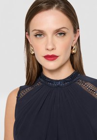 Námornícka sukňa s plisovaným materiálom a priesvitnými čipkovými detailmi na pleciach. Doplnkovaná korálkovým golierom a v kombinácii so zlatými kruhovými náušnicami.