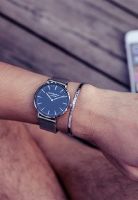 Montre en acier inoxydable ronde avec un cadran bleu foncé, des aiguilles argentées et un bracelet en maille. Accompagnée d'un bracelet en argent avec un texte gravé.