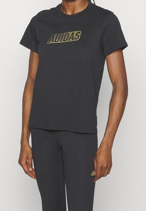 Personne portant un t-shirt noir Adidas avec un logo à rayures jaunes et un pantalon noir avec un petit logo Adidas jaune sur la cuisse.