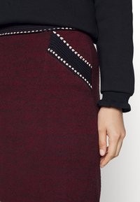 Bordó pletená sukně s černou tkanou kapsou a diagonálními stehy jako akcenty. Hladká textura; pohodlné nošení. Ruka spočívá na sukni.