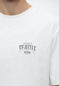 Λευκό μπλουζάκι με το κείμενο "The Adventure City SEATTLE USA" εκτυπωμένο στο αριστερό στήθος, φορεμένο από άτομο με περικοπή στο λαιμό.