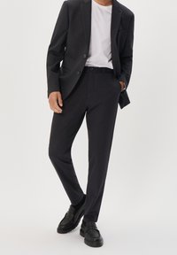 Donkergrijs pak met een op maat gemaakte jas en bijpassende broek, gecombineerd met een wit T-shirt en zwarte loafers. Gladde textuur, strakke lijnen.