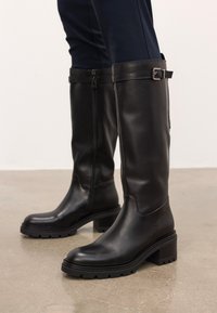 Egy nő fekete, térdig érő bőr csizmát visel, amely blokksarkú és oldalsó cipzárral rendelkezik, sima betonpadlón áll.