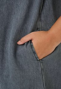 Grått tygplagg med en texturerad yta, med en sidoficka. En hand vilar i fickan och visar plaggdesignen.