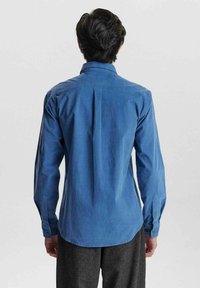 Camisa de manga larga azul de tela de algodón, con un pliegue en la parte posterior, puños abotonados y un dobladillo curvedo. Textura suave y diseño de cuello clásico.