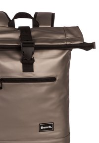 Sac à dos gris à rabat, fabriqué en matériau durable, avec des zips noirs, des sangles ajustables et une poche avant zippée. Logo de la marque affiché de manière proéminente.