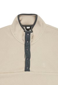 Sudadera de forro polar en blanco roto con un cuello alto. Presenta una tapeta de botones en gris oscuro y un logotipo bordado en la parte frontal. Textura suave.