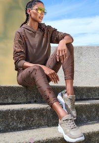 Brauner Kapuzenpullover mit grafischen Akzenten, passende braune Leggings mit Wirbelmustern und graue Sneakers mit weißen Sohlen. Reflektierende Sonnenbrille.