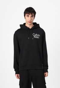 Black hooded sweatshirt made of cotton, featuring a front pocket and a white "Calvin Klein" logo on the left side. Smooth texture.