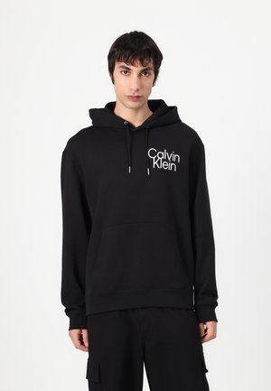 Zwarte hoodie van katoen, met een voorzak en een wit "Calvin Klein" logo aan de linkerkant. Glad textuur.