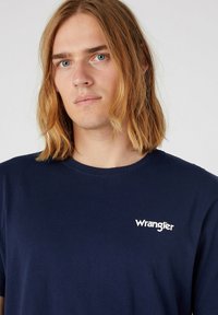 Ung person med skulderlangt blond hår iført en navyblå Wrangler T-skjorte mot en ensfarget lys bakgrunn.
