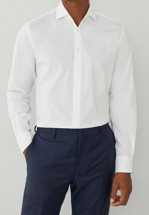 Homme portant une chemise blanche cintrée boutonnée rentrée dans un pantalon de costume bleu foncé, une main dans la poche.