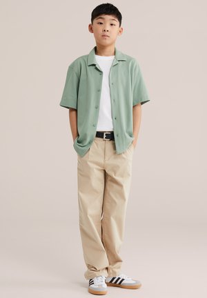 Junge steht mit Händen in den Taschen, trägt ein hellgrünes Kurzarmhemd über einem weißen T-Shirt, beige Hose und weiße Turnschuhe mit schwarzen Streifen.