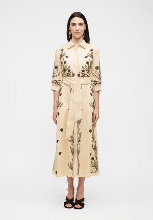 Kvinde med langt mørkt hår iført en beige midi-kjole med mørkt botanisk broderi, 3/4-ærmer, bælte i taljen og sorte åbne tå-hæle.