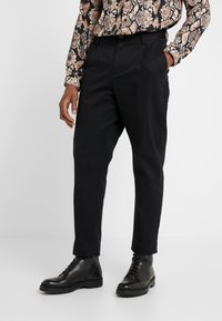 Černé zúžené kalhoty s hladkým povrchem, mají plochý přední díl a elastický pas, doplněné černými koženými botami.