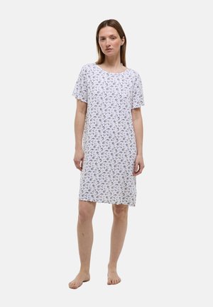 Femme debout pieds nus portant une chemise de nuit blanche jusqu’aux genoux, à manches courtes, avec un petit imprimé floral sur un fond uni.