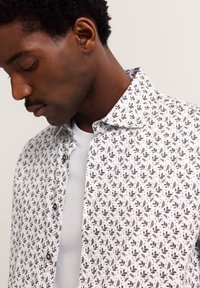 Mann mit geschlossenen Augen, der ein weißes Hemd mit kleinen grünen und braunen Blattmustern über einem schlichten weißen T-Shirt trägt.